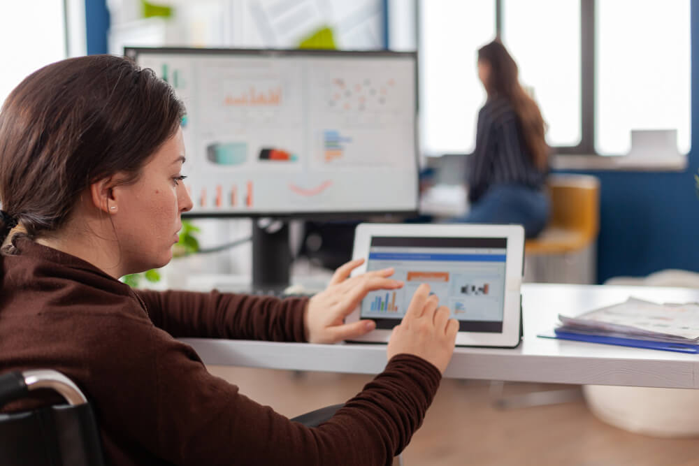 mulher à mesa do escritorio, usando computador e tablet ao mesmo tempo, verificando dados estatísticos